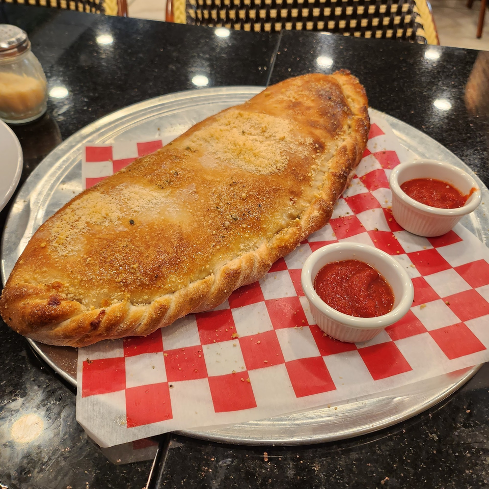 Golden-brown calzone with parmesan cheese, served on a metal tray with two cups of marinara sauce.